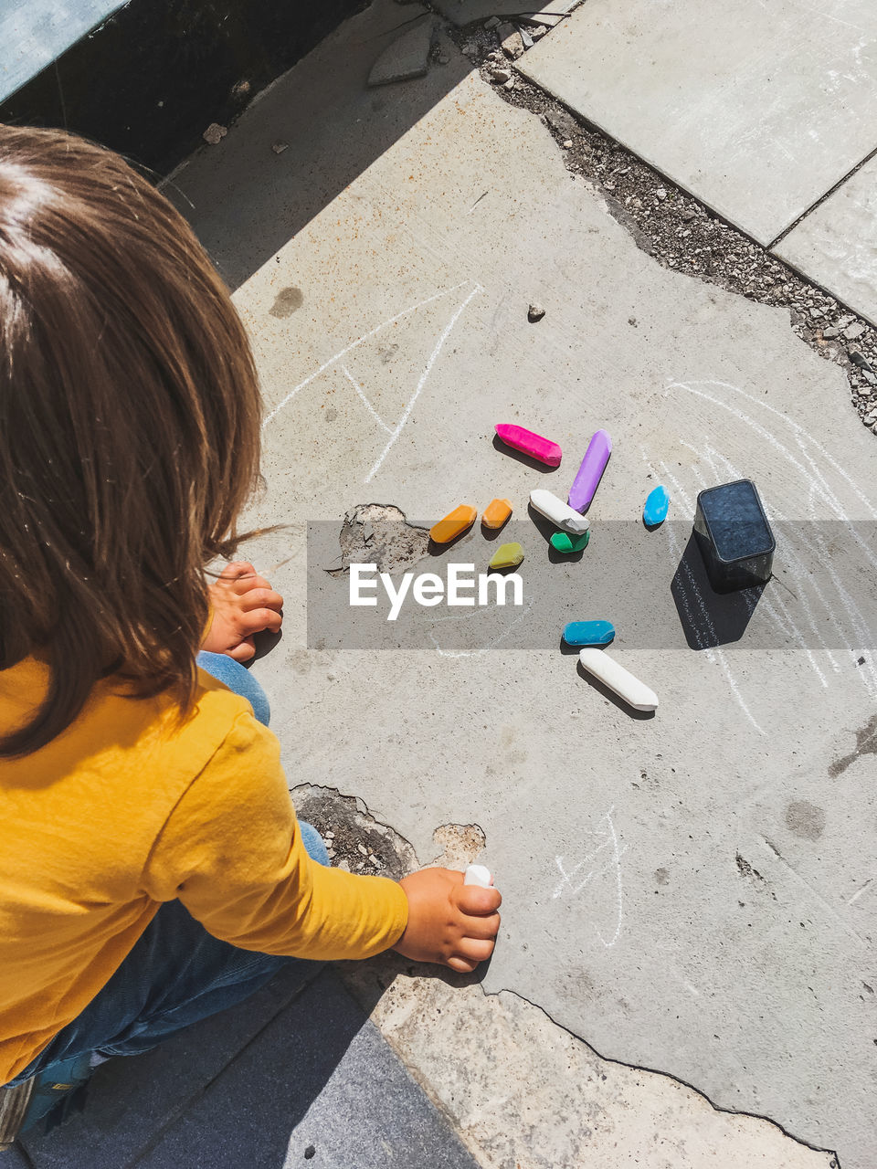 Toddler in jeans draws with crayons on the asphalt in sunny day. kid's hands are covered with stain. 