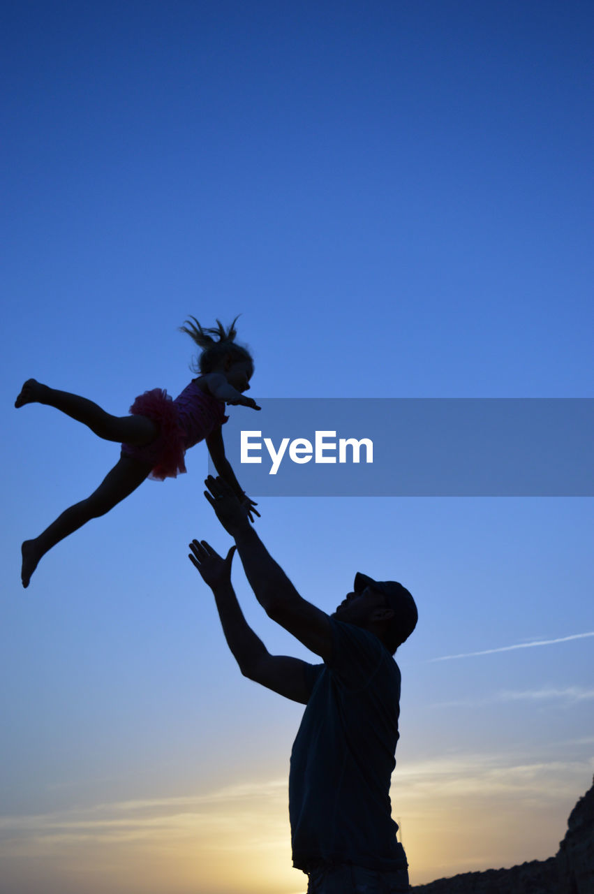 Low angle view of silhouette man catching daughter against blue sky during sunset