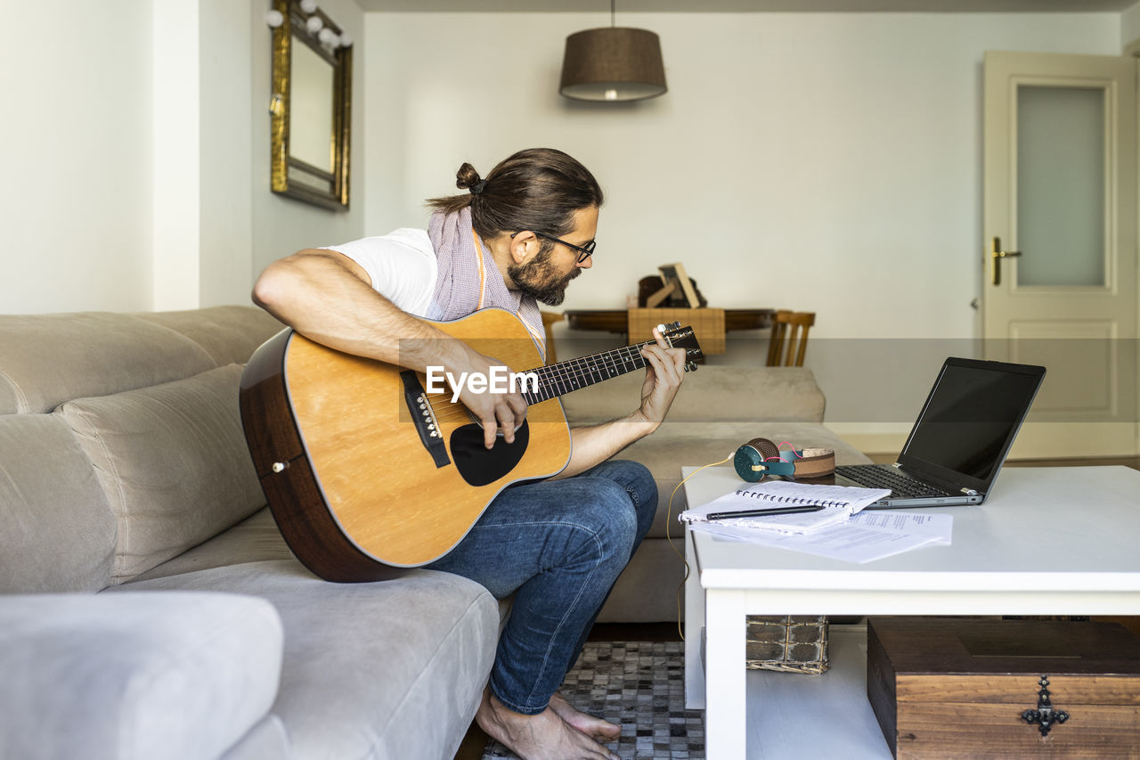 Side view of bearded creative male musician in casual wear sitting on sofa and playing acoustic guitar in modern living room