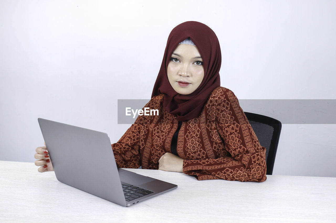 PORTRAIT OF YOUNG WOMAN USING SMART PHONE WHILE SITTING ON LAPTOP
