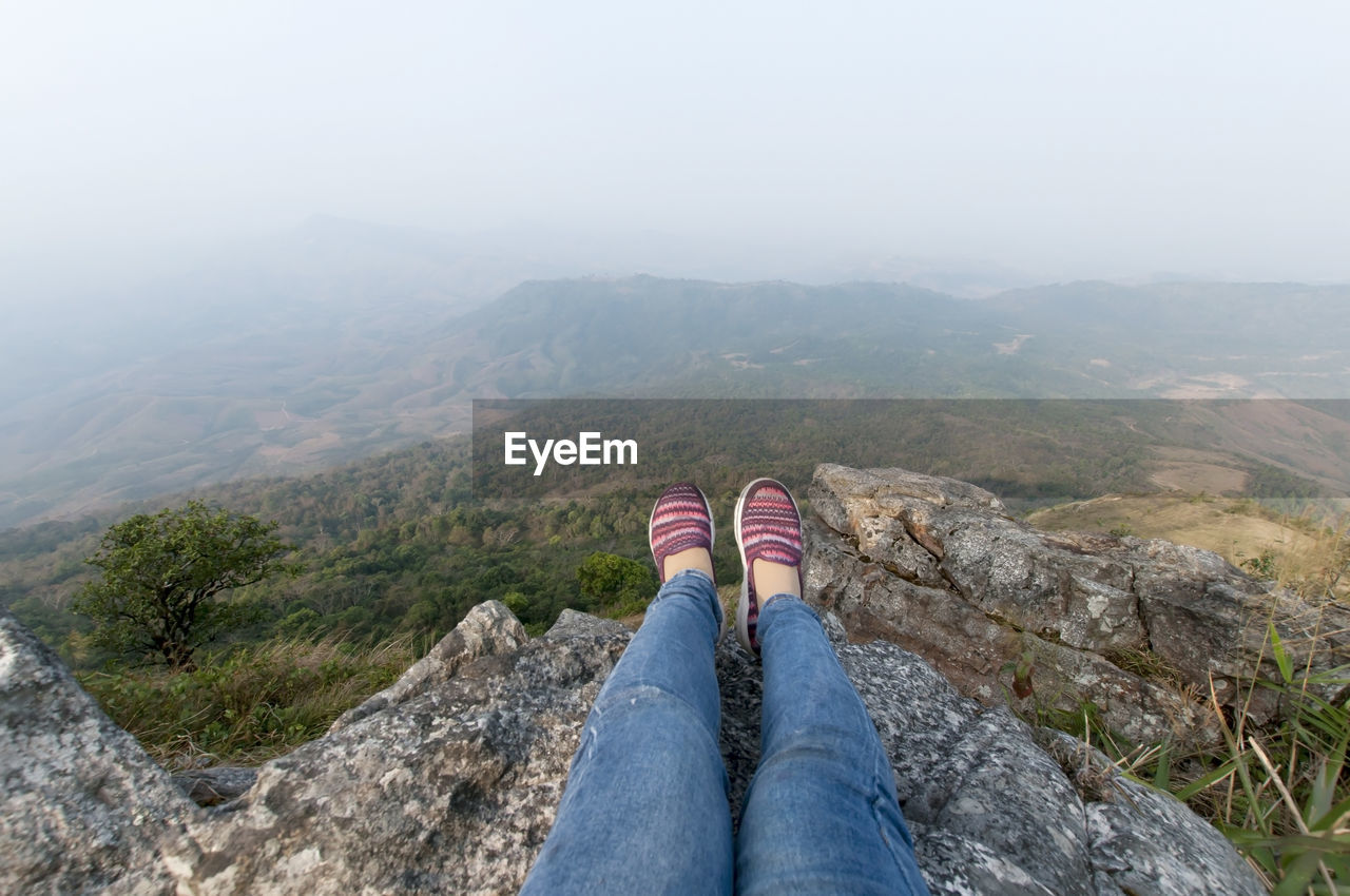 Low section of woman sitting on rock formation over mountains in foggy weather