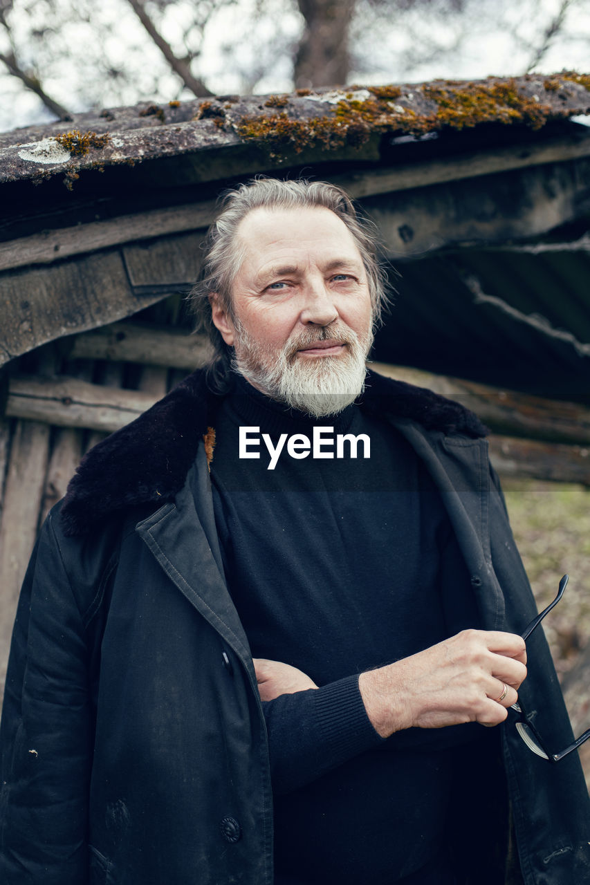 Elderly man with a beard stands in the village near a wooden shed in a sheepskin coat and glasses