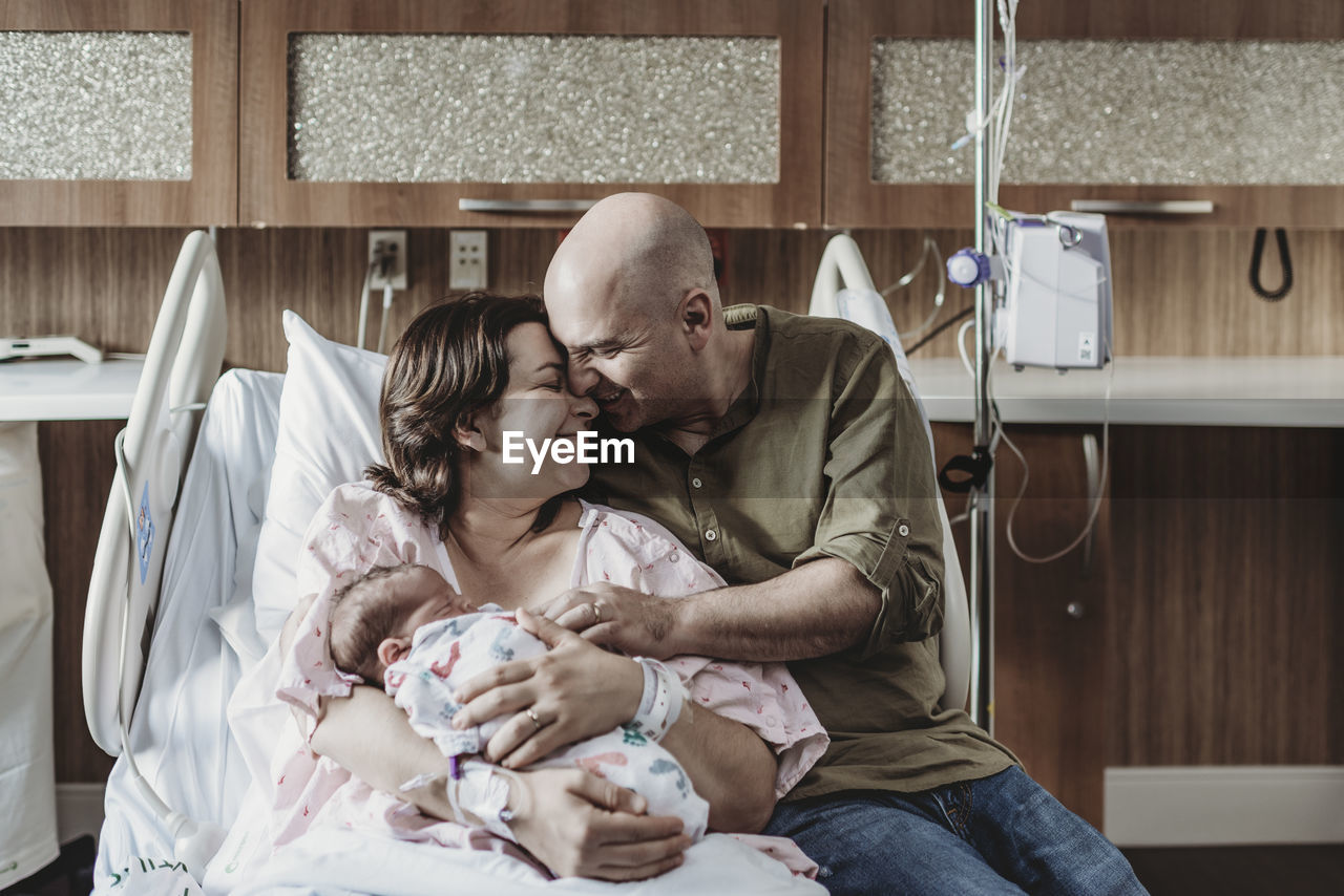 Portrait of father embracing newborn and wife in hospital bed