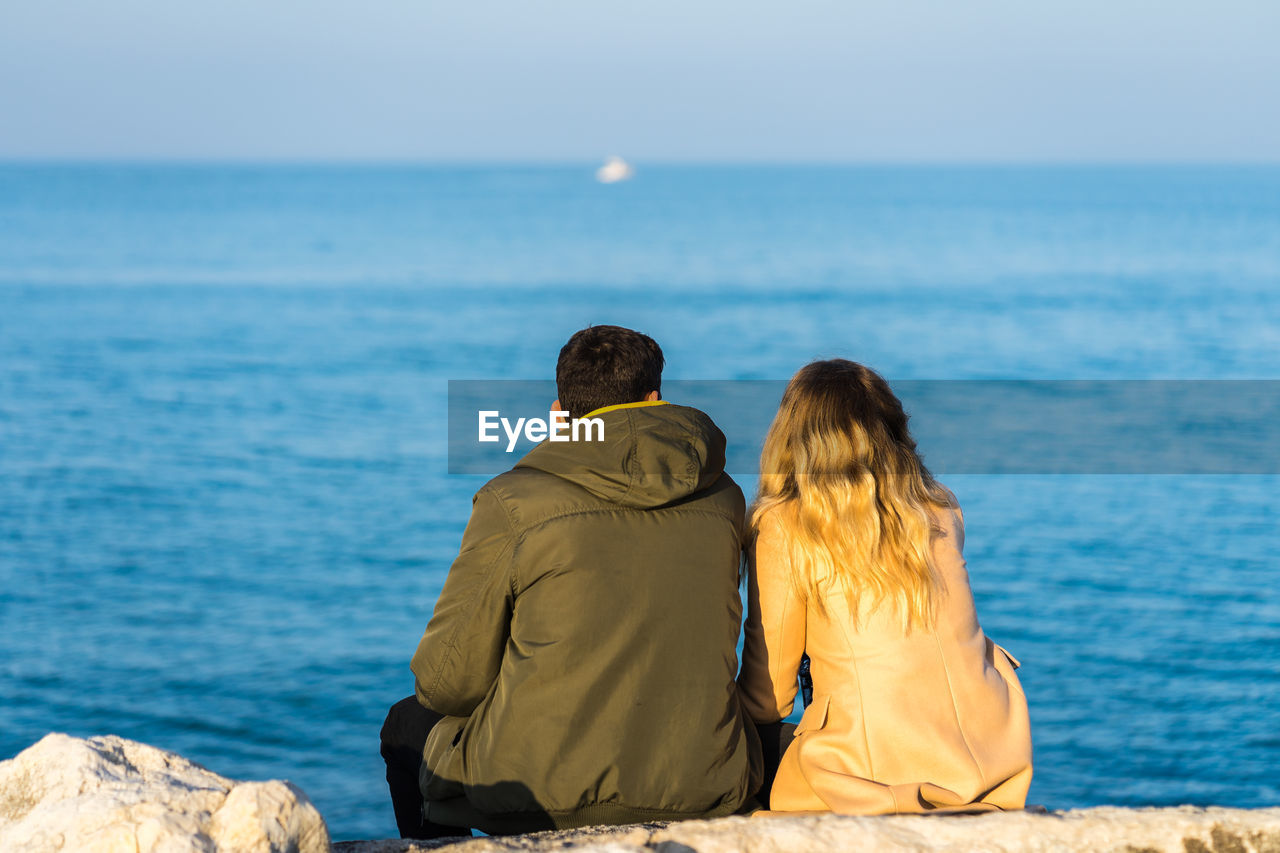 REAR VIEW OF COUPLE SITTING ON SHORE AGAINST SEA
