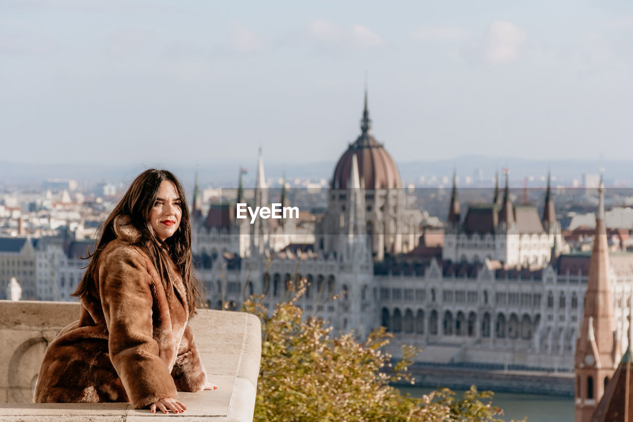 Portrait of beautiful young woman on balcony overlooking hungarian parliament in budapest, hungary