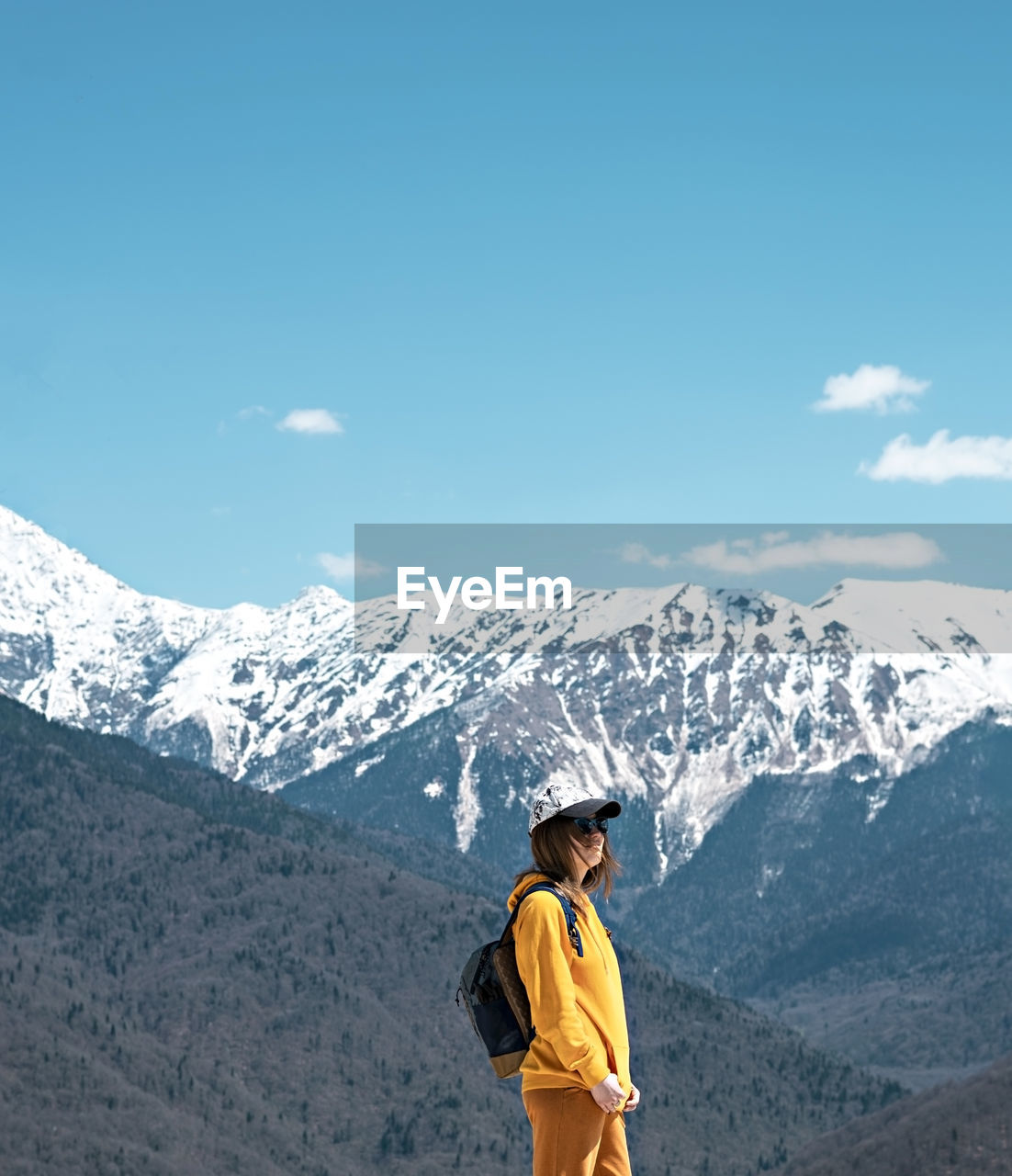 Young blond woman in yellow hoodie, cap, on background of snow capped caucasus mountains. hiking