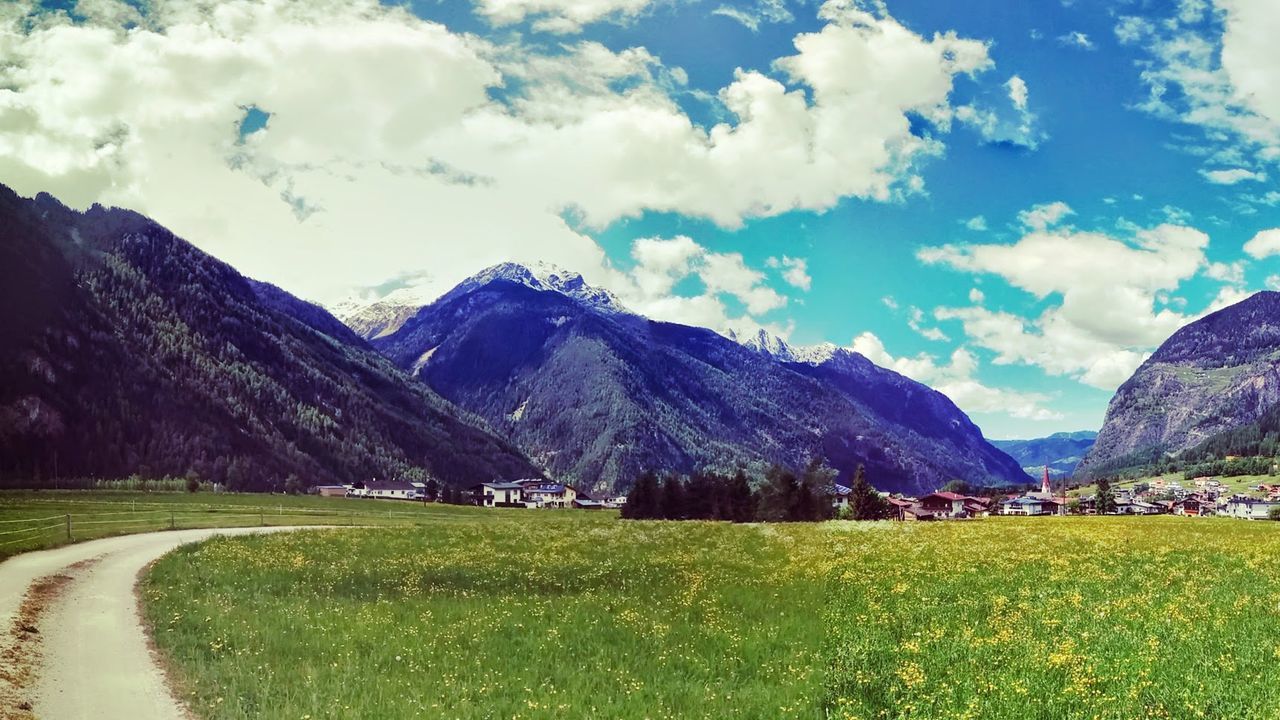 Dirt road by grassy field against mountains and cloudy sky