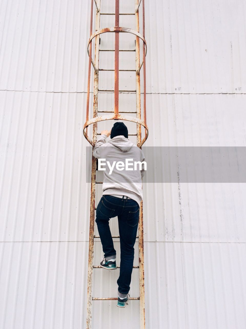 Rear view full length of man climbing on rusty ladder by white wall