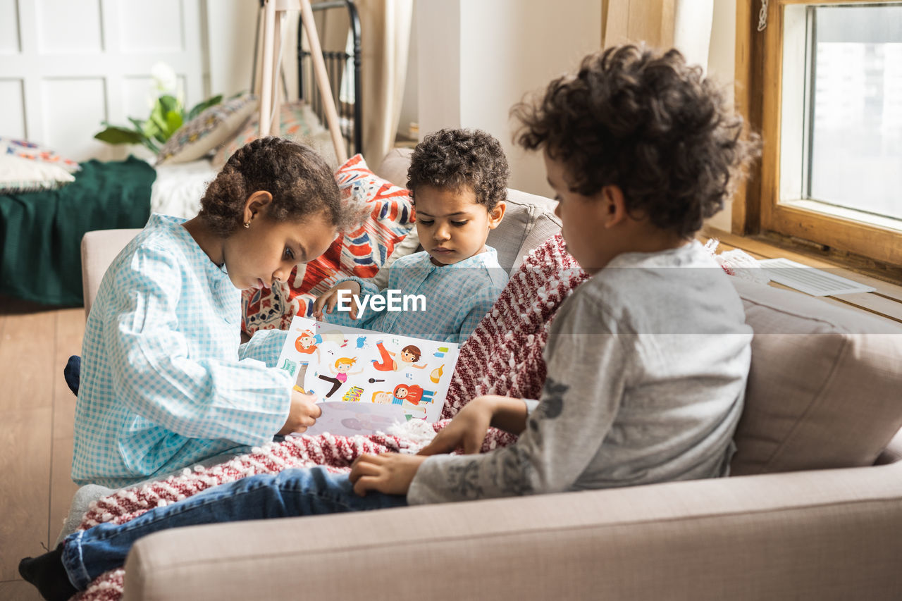 Siblings with book on sofa at home