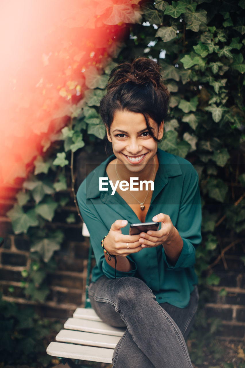 Portrait of young woman using mobile phone while sitting on chair in backyard