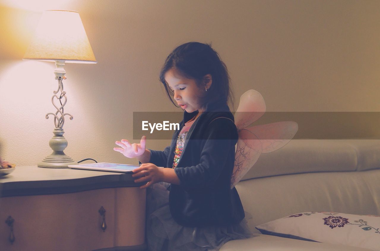 Girl wearing fairy wings using digital tablet while sitting on sofa by illuminated lamp shade