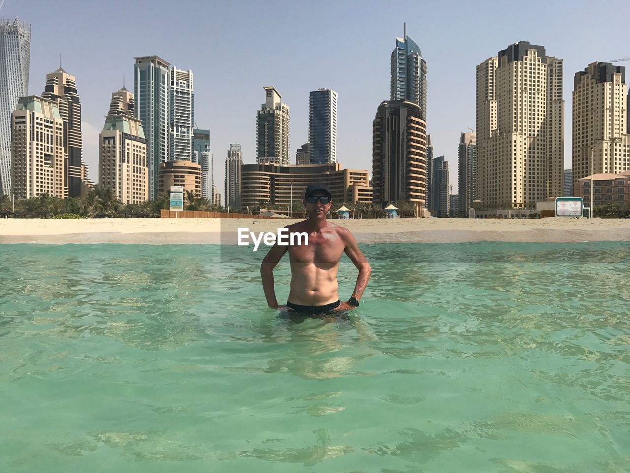 Portrait of shirtless man standing at beach against modern buildings in city