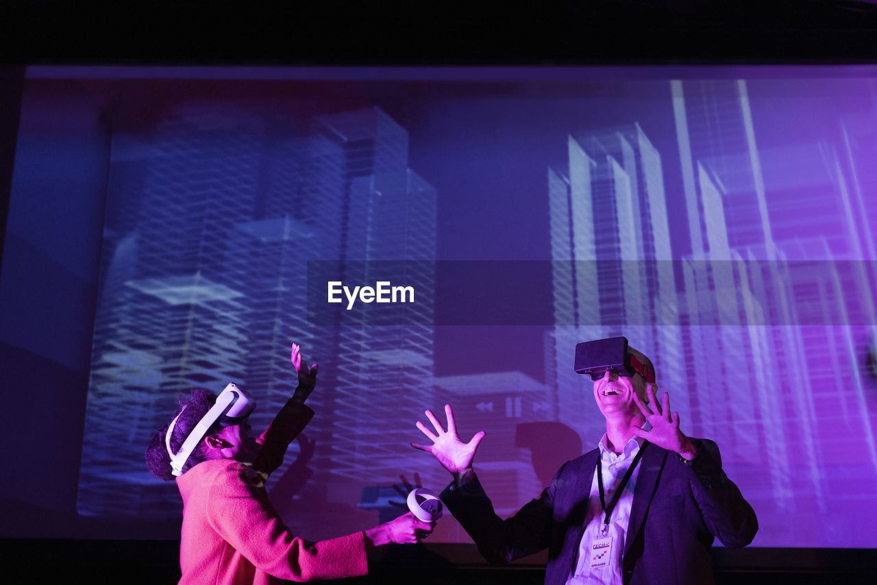 Happy male and female entrepreneurs with futuristic glasses gesturing by projection screen at convention center
