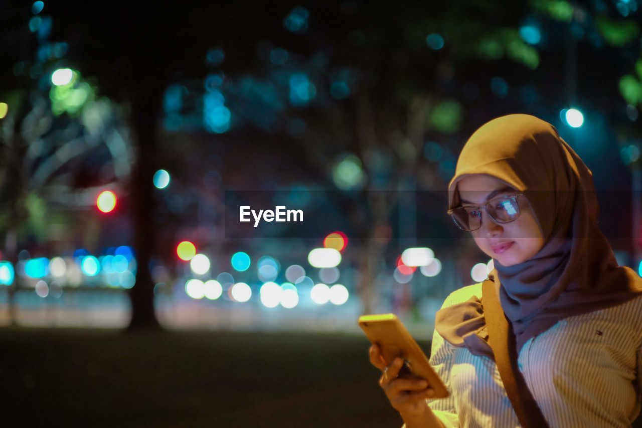 Woman wearing hijab using phone outdoors at night
