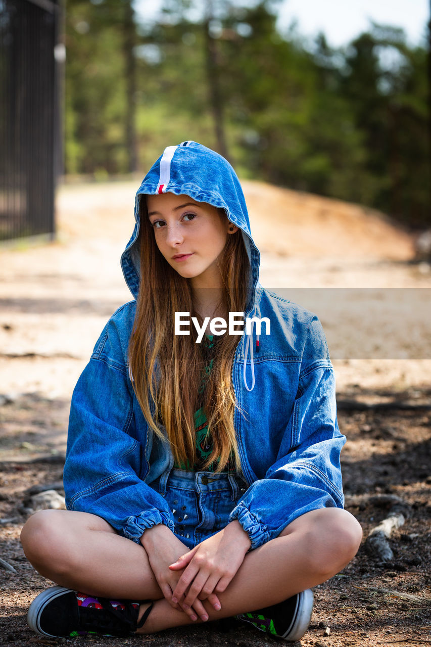 A young girl poses in a denim jacket with a hood against the background of a forest sitting
