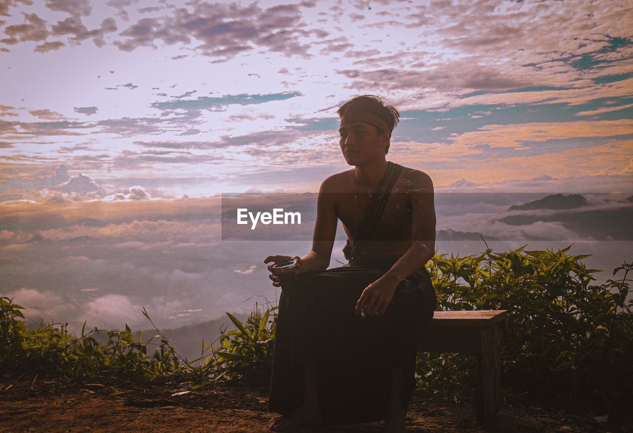 Young man holding drink sitting against sky during sunset