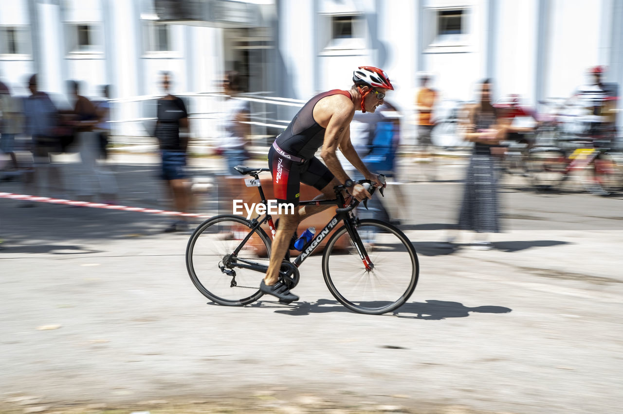 bicycle, sports, transportation, motion, activity, blurred motion, cycling, city, road bicycle, architecture, speed, endurance sports, adult, road cycling, vehicle, lifestyles, racing bicycle, city life, street, sports equipment, men, city street, exercising, mode of transportation, riding, competition, wheel, athlete, bicycle racing, headwear, commuter, recreation, muscular build, helmet, copy space, clothing, road bicycle racing, race, building exterior, cycle sport, road, sports race, person, bicycle wheel, full length, one person, vitality, built structure, on the move, bicycle helmet, sports clothing, competitive sport