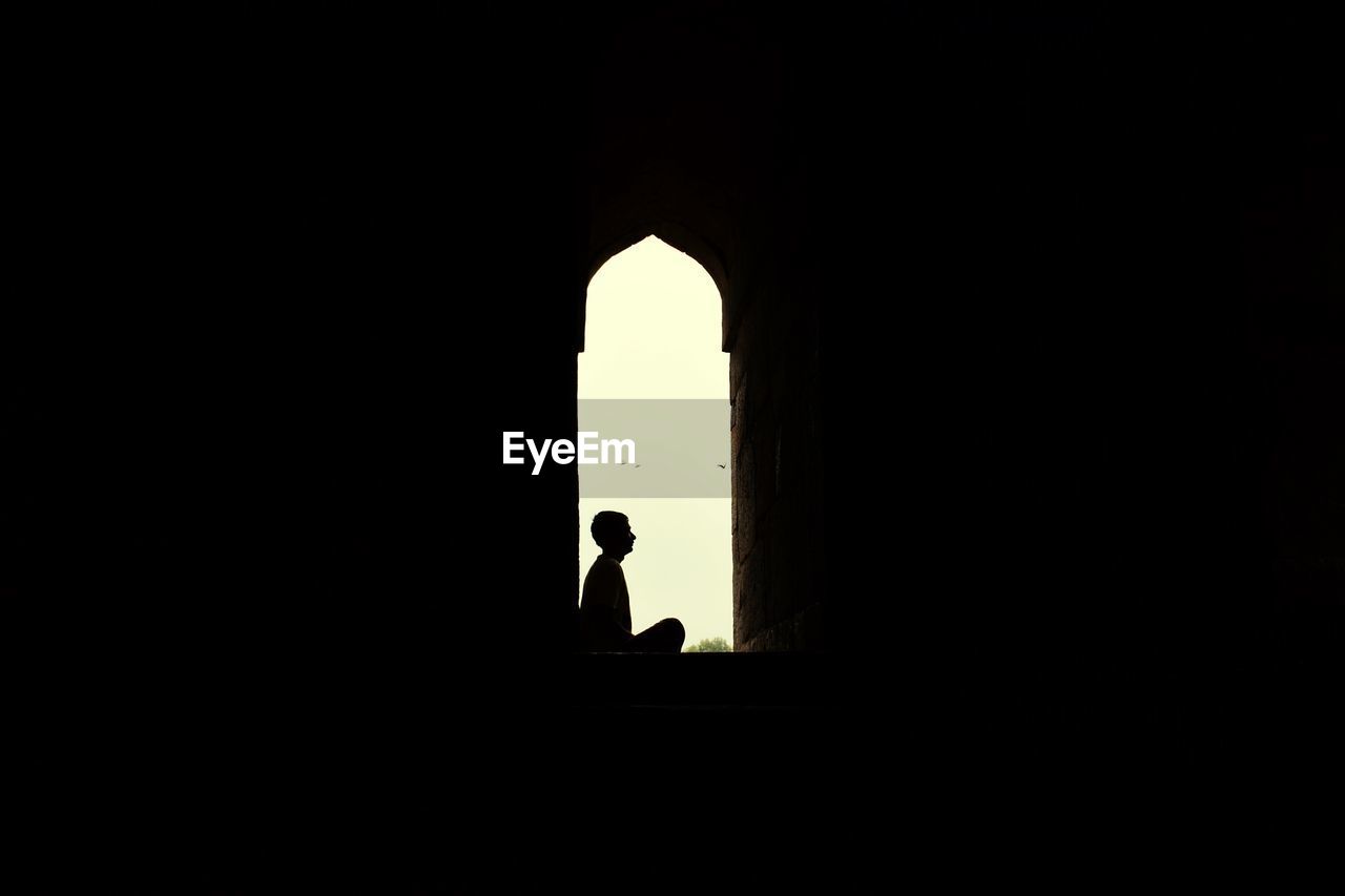 Silhouette man sitting on window sill against clear sky