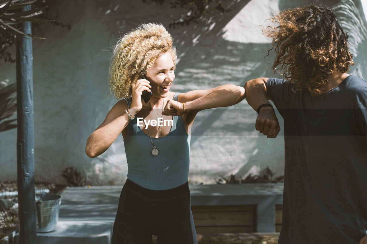 Smiling woman giving elbow bump to man while talking on mobile phone outdoors