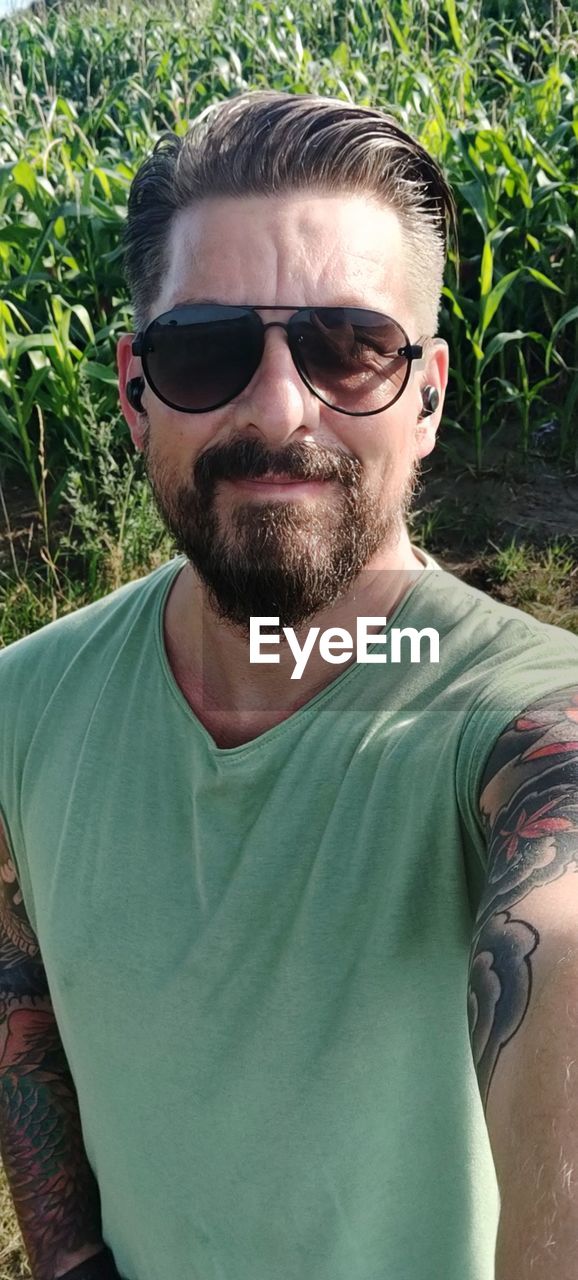 glasses, sunglasses, fashion, one person, portrait, beard, facial hair, adult, human hair, men, front view, hairstyle, looking at camera, young adult, casual clothing, leisure activity, lifestyles, green, plant, person, nature, t-shirt, day, headshot, waist up, tattoo, goggles, cool attitude, outdoors, grass, clothing, sunlight, eyeglasses, human face, smiling, standing, stubble, vision care