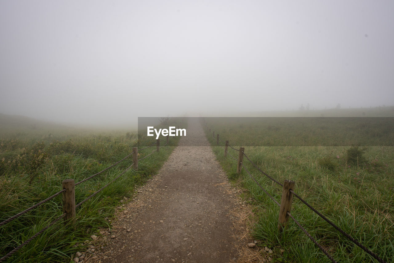 Dirt road amidst field against sky during foggy weather