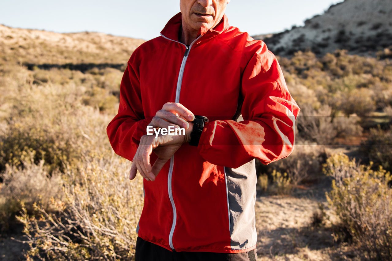 Concentrated middle aged male athlete in activewear checking time on smartwatch while standing in hilly terrain after workout