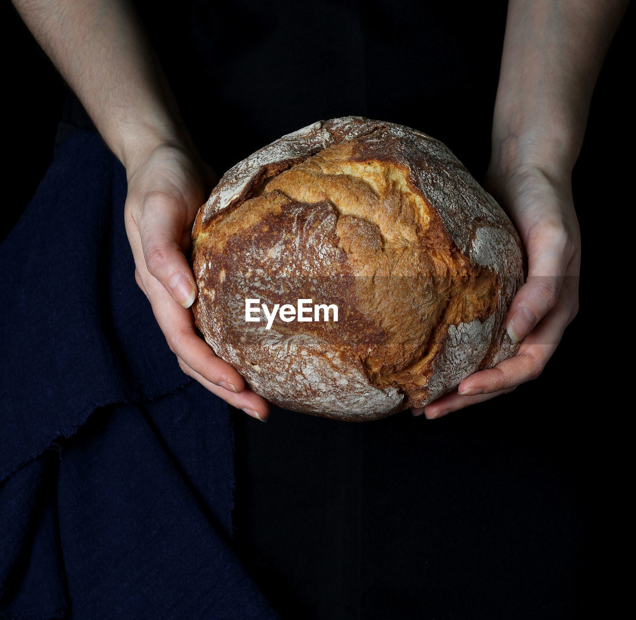 Round rustic bread held with the hands, above the black backdrop with the canvas on the left side