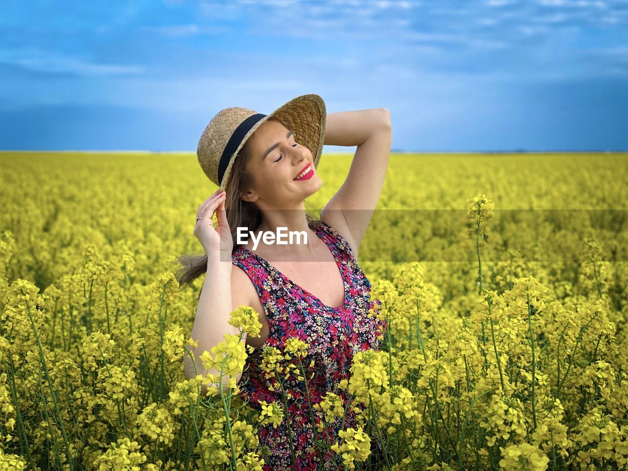 Portrait of young woman wearing dress and sun hat in a field of canola flowers on a cloudy day