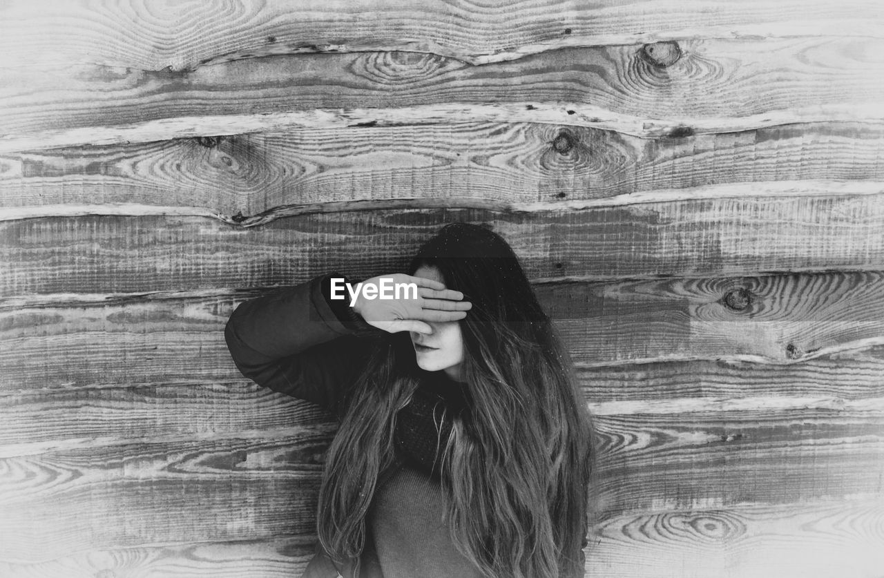 Young woman covering eyes against wooden wall