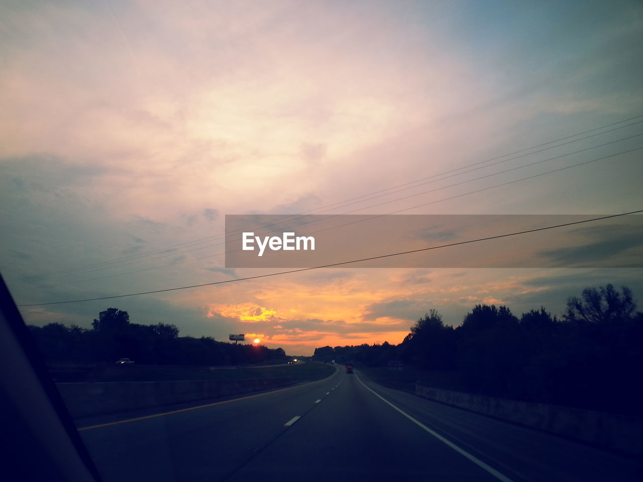 Silhouette trees amidst road against sky during sunset seen from car windshield