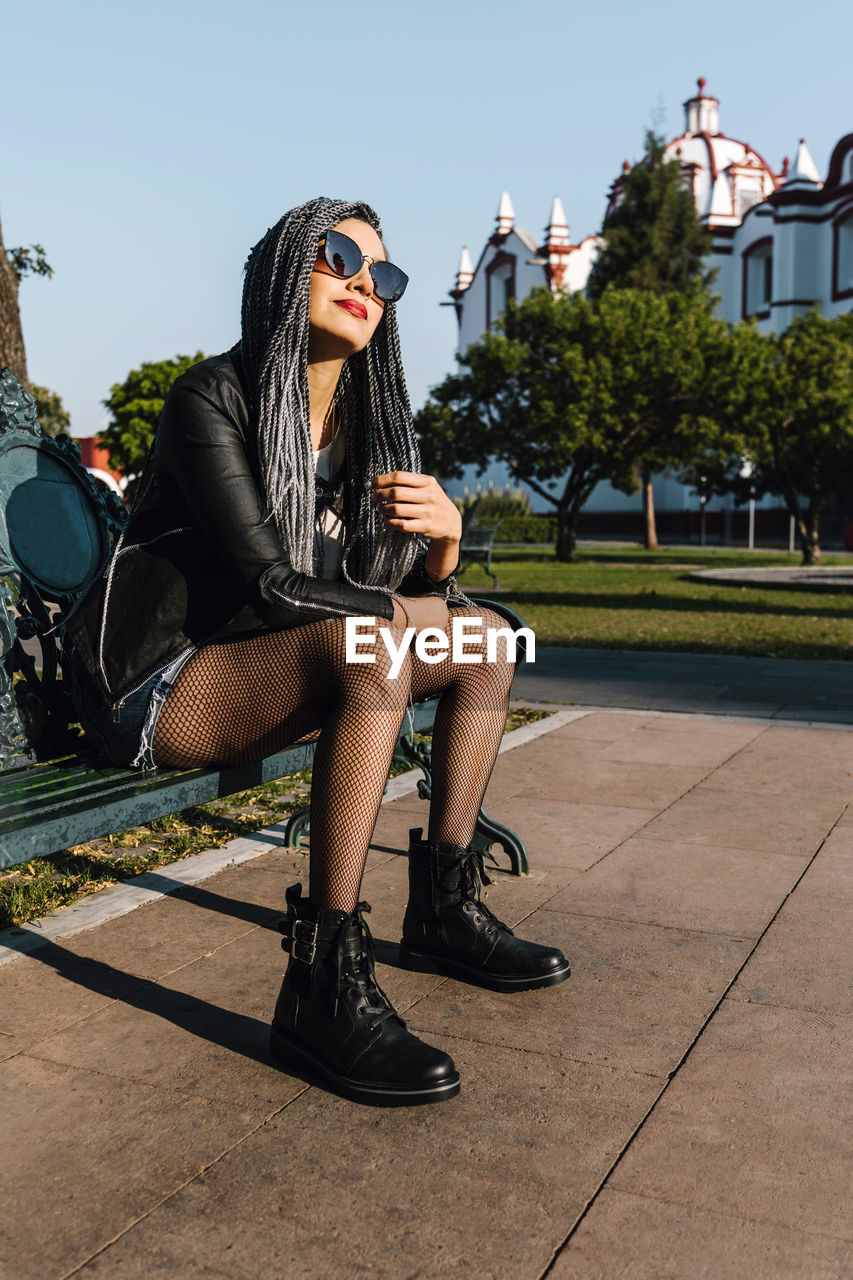 Cool latin american female with afro braids in modern sunglasses and leather boots sitting on bench in sunny park