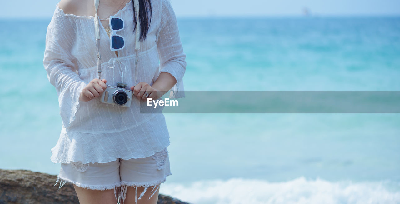 Midsection of woman with camera standing against sea