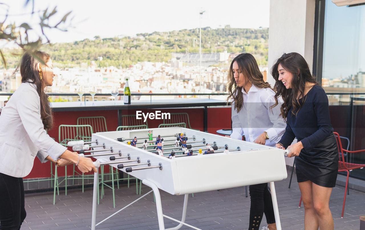 Happy adult hispanic girlfriends in smart casual clothes smiling and playing table football together during party on weekend day on terrace