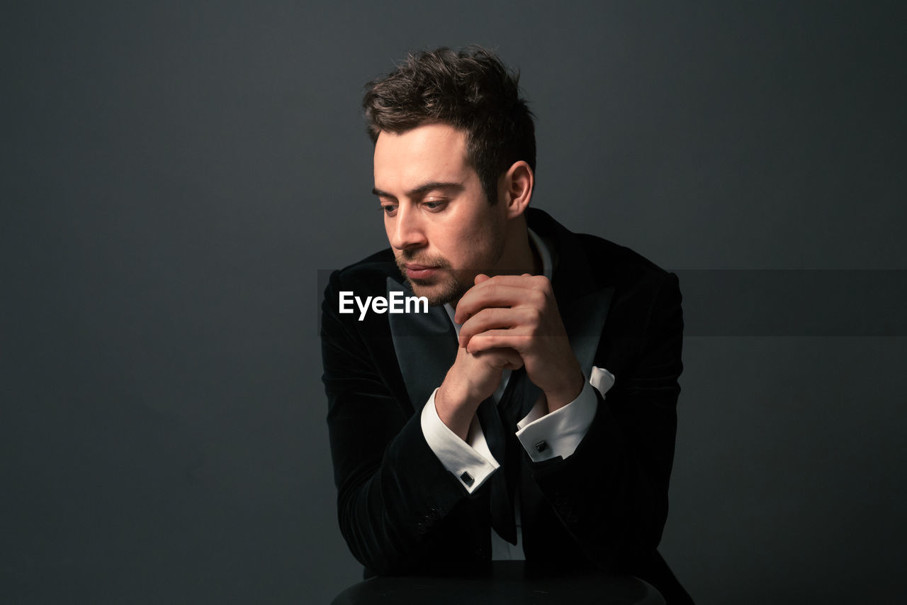 Thoughtful young man in suit sitting with hands clasped against gray background