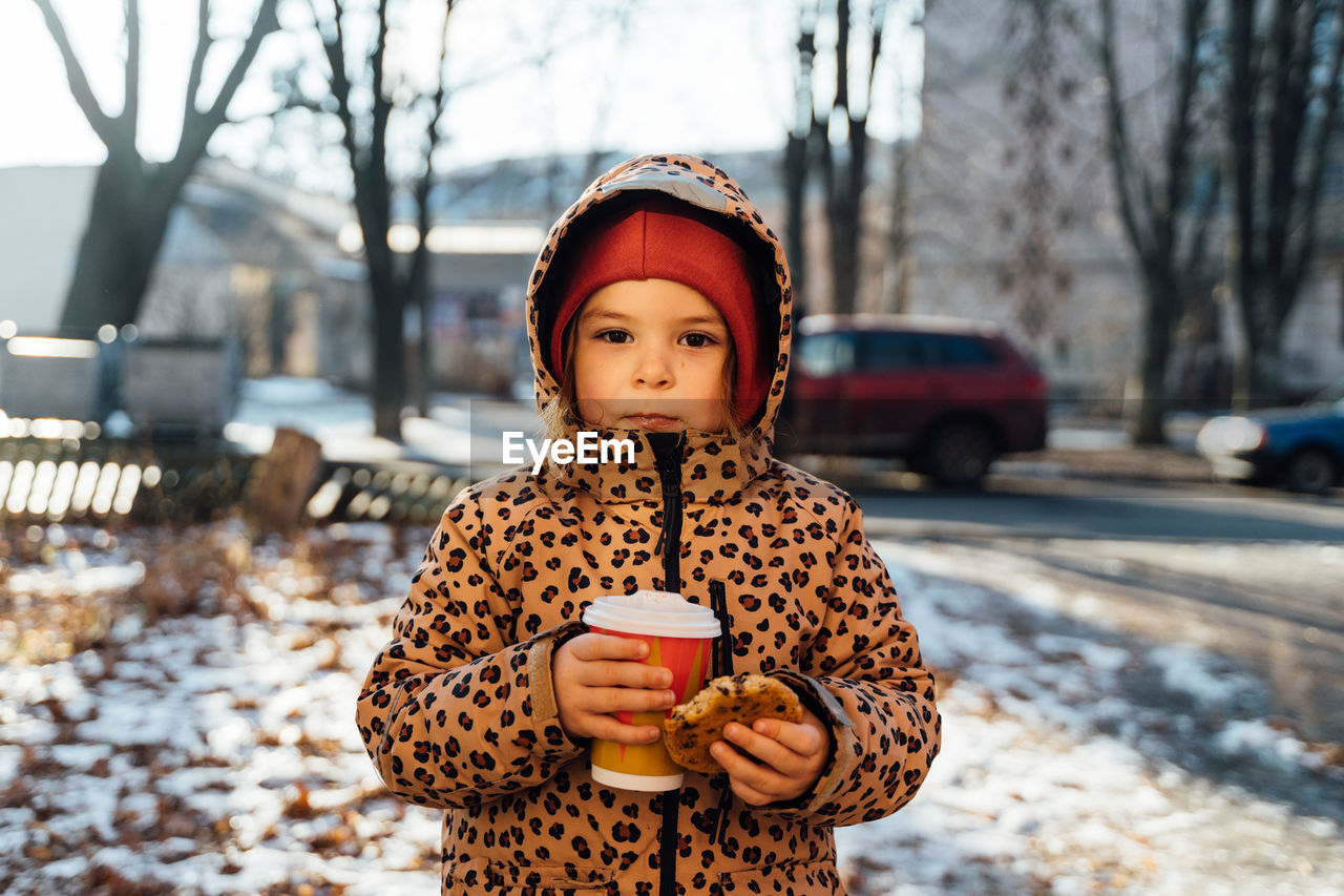 Cute girl in warm clothes holding cookie and cup of takeaway drink while looking at camera in the street in winter day