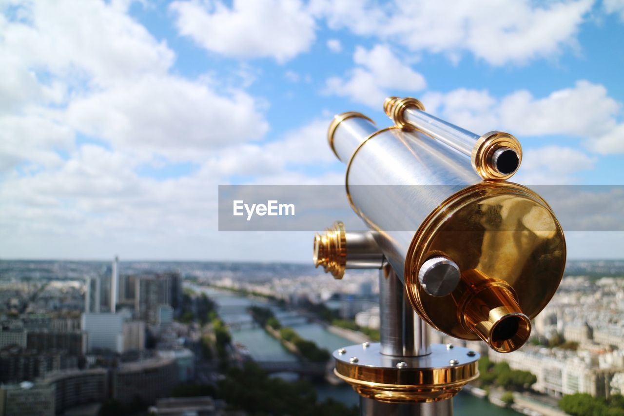 Close-up of coin-operated binoculars with cityscape in background against sky