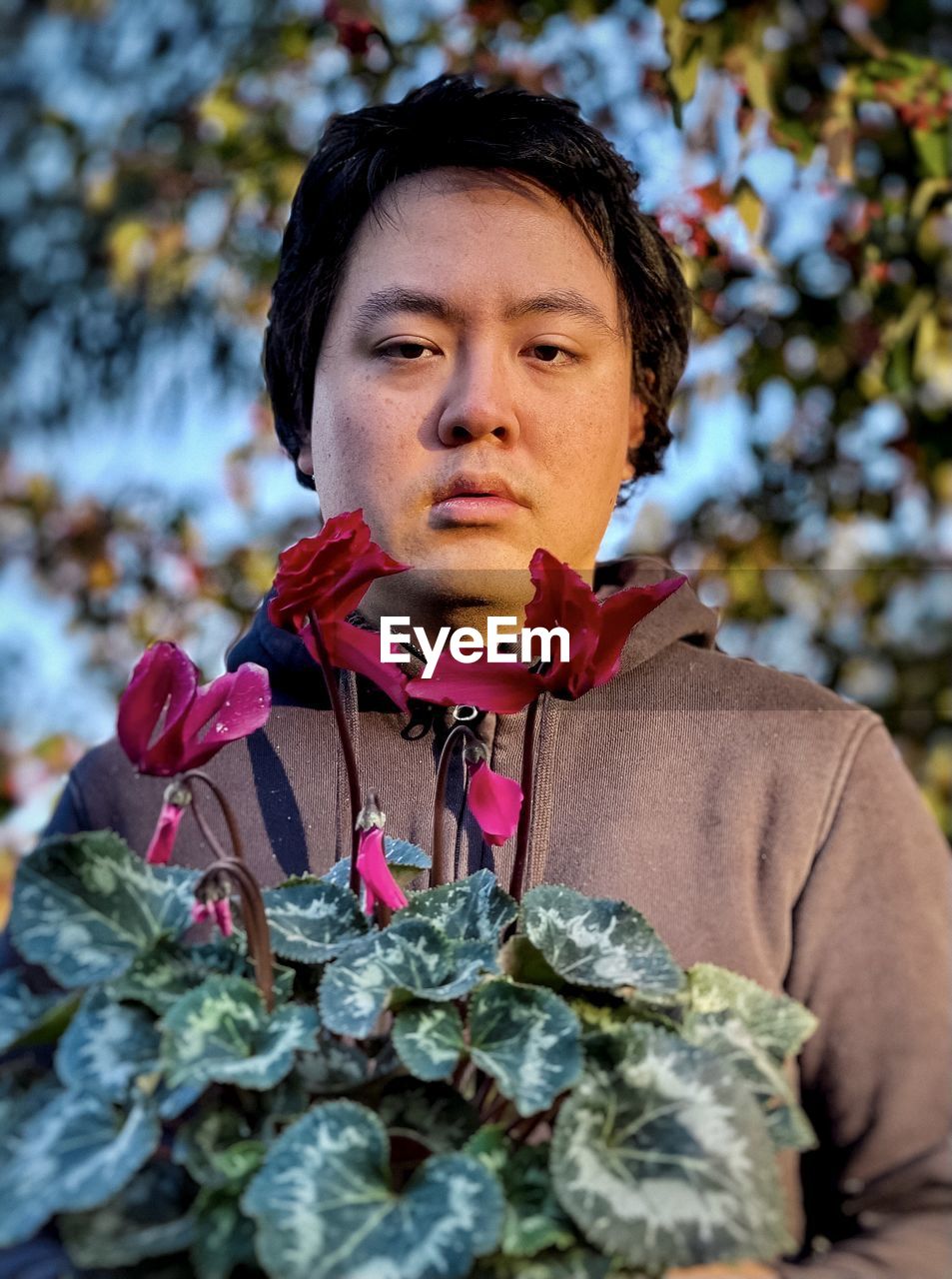 Portrait of young asian man with red flowering cyclamen plant against trees.