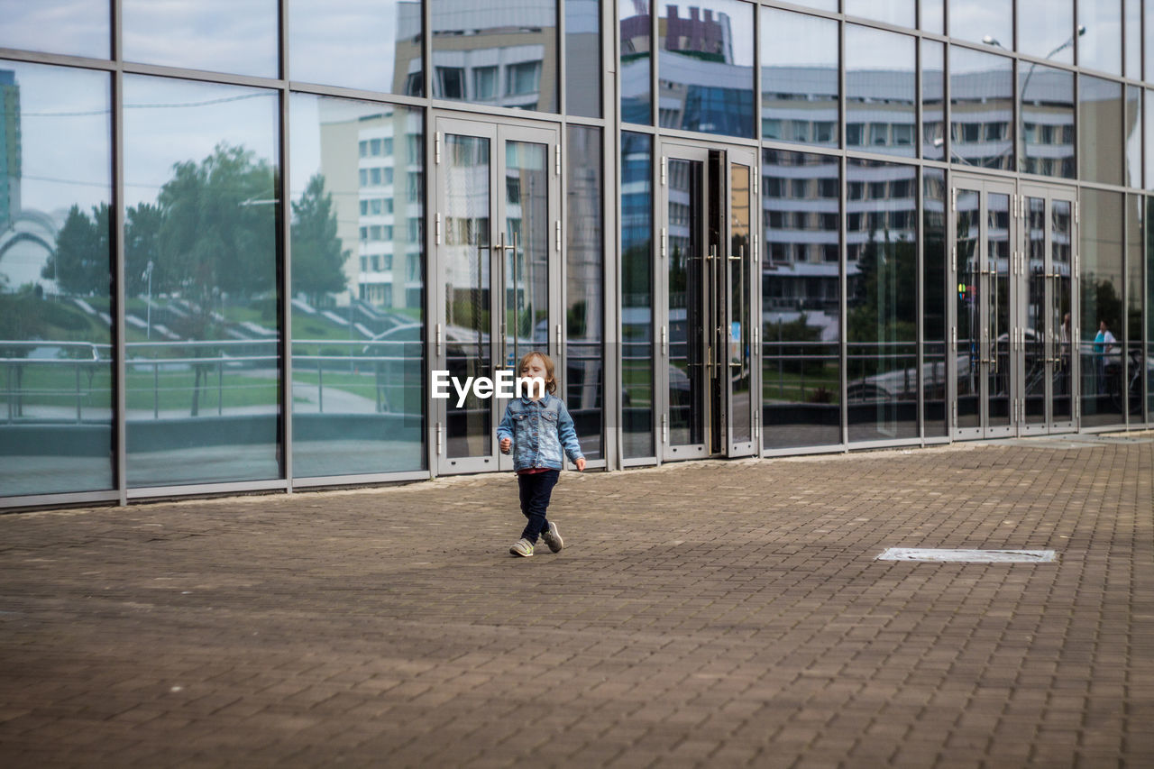 Girl walking on footpath by glass building