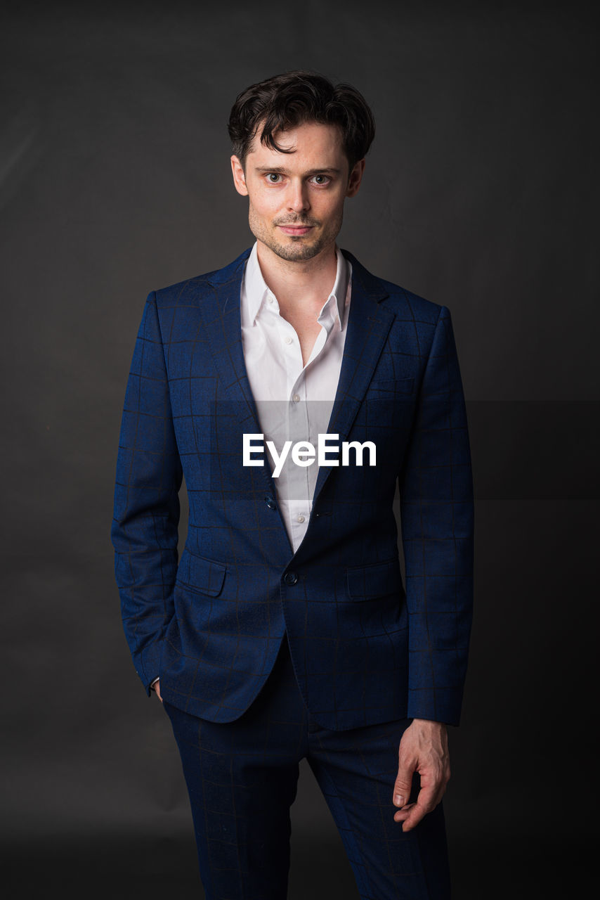 one person, adult, portrait, formal wear, business, men, studio shot, looking at camera, tuxedo, indoors, blazer, front view, businessman, young adult, standing, clothing, gray, fashion, brown hair, three quarter length, hands in pockets, person, business finance and industry, serious, photo shoot, elegance, button down shirt, occupation, gray background, individuality, corporate business, spring, success, looking