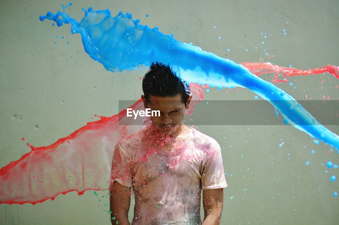 Color water splashing over young man standing against gray background