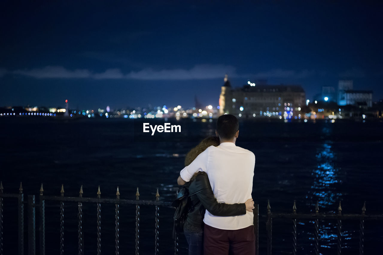 Rear view of man embracing woman while standing against sea at night