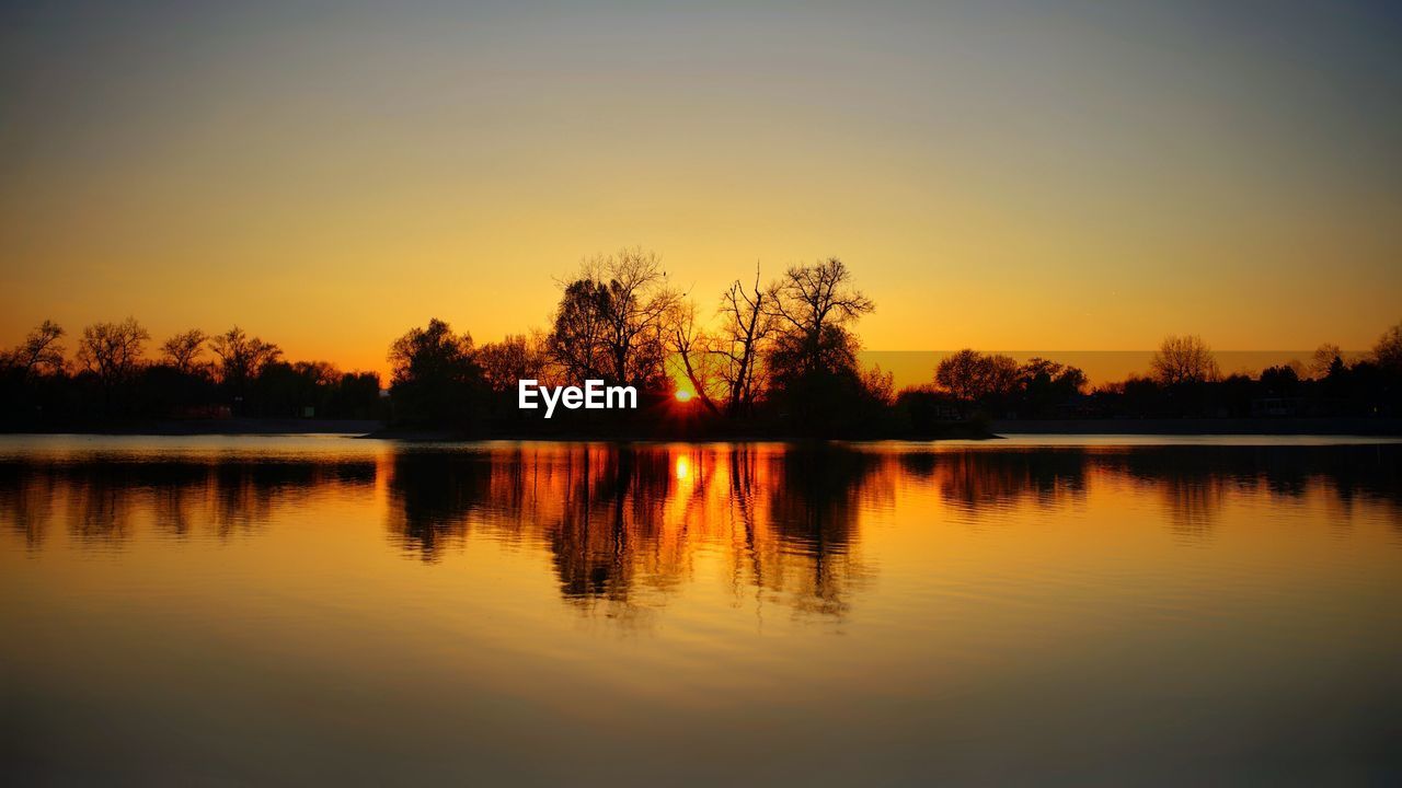 REFLECTION OF SILHOUETTE TREES IN LAKE AGAINST SKY DURING SUNSET