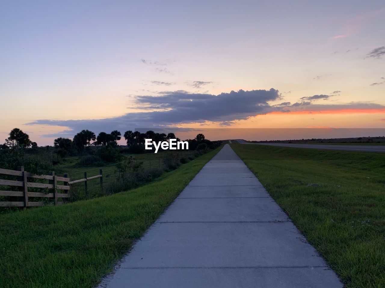 Footpath amidst field against sky during sunset