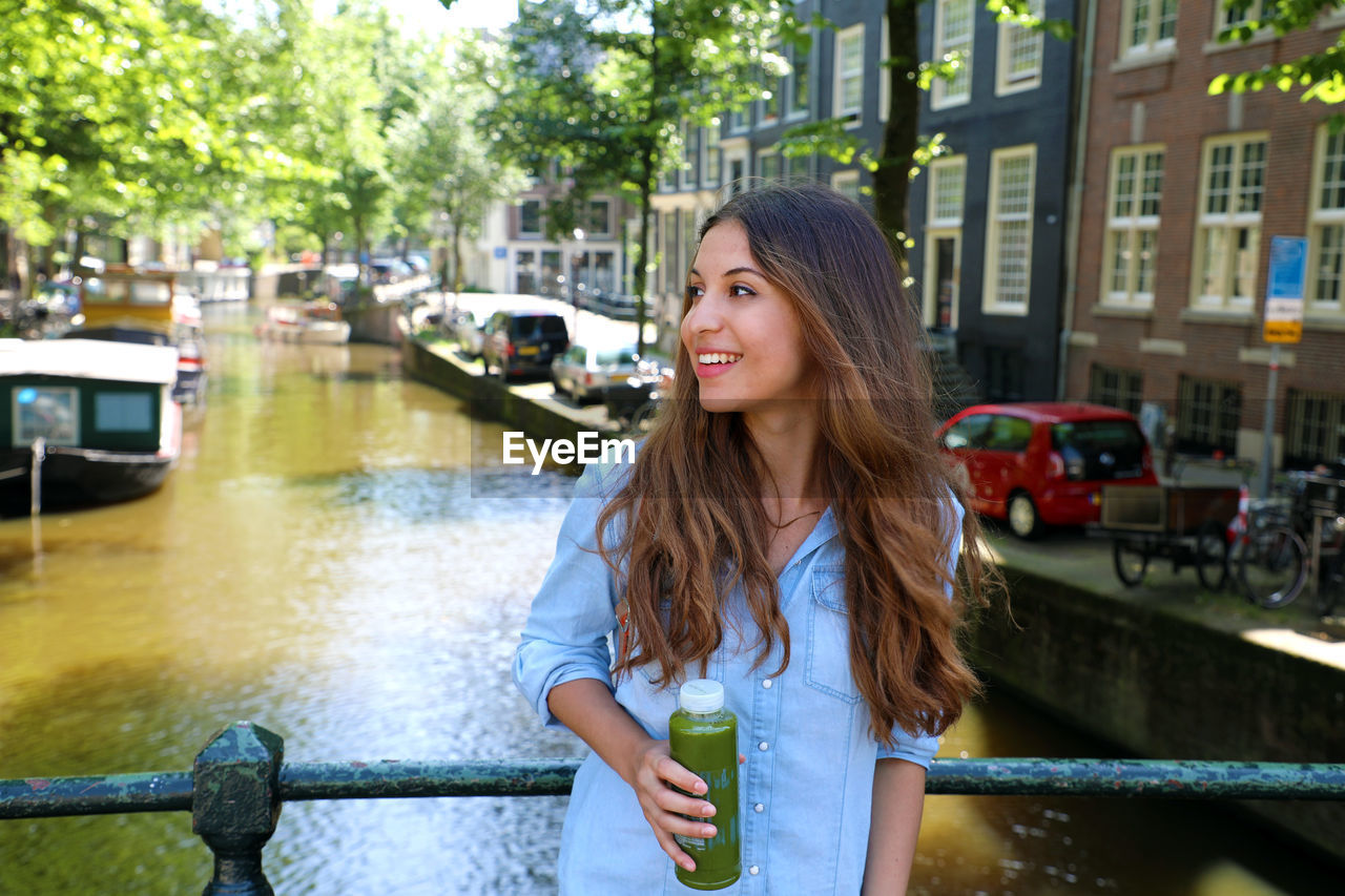 BEAUTIFUL WOMAN SMILING WHILE STANDING BY RAILING IN CANAL