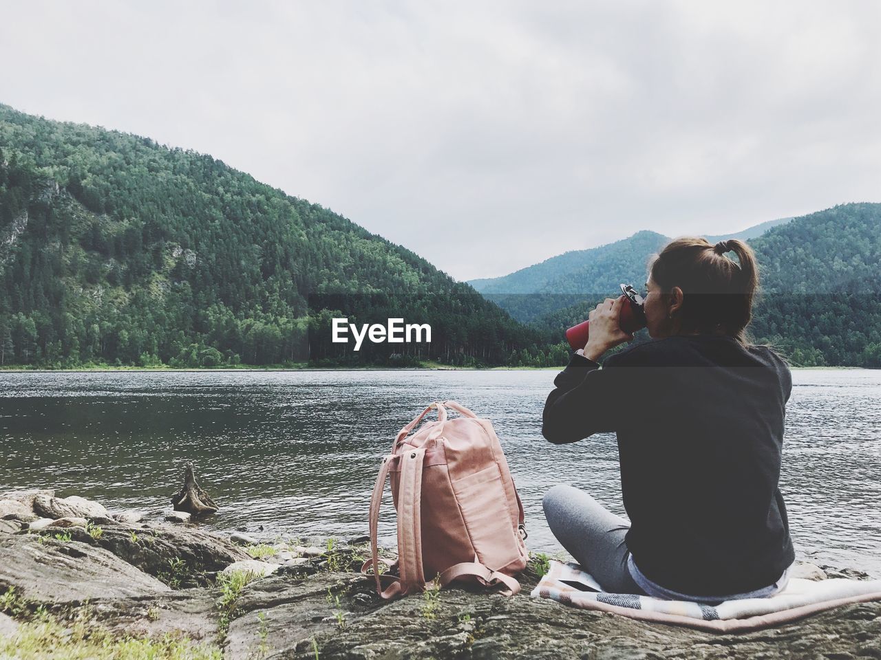 Rear view of woman drinking water while sitting at lake against mountain