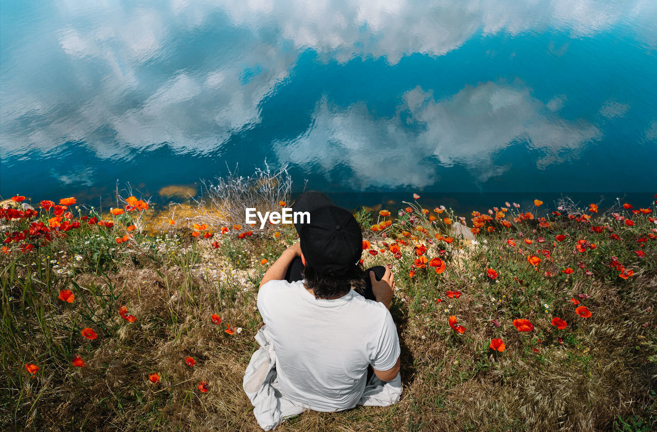 Man sitting in poppy field by the lake with clouds reflection