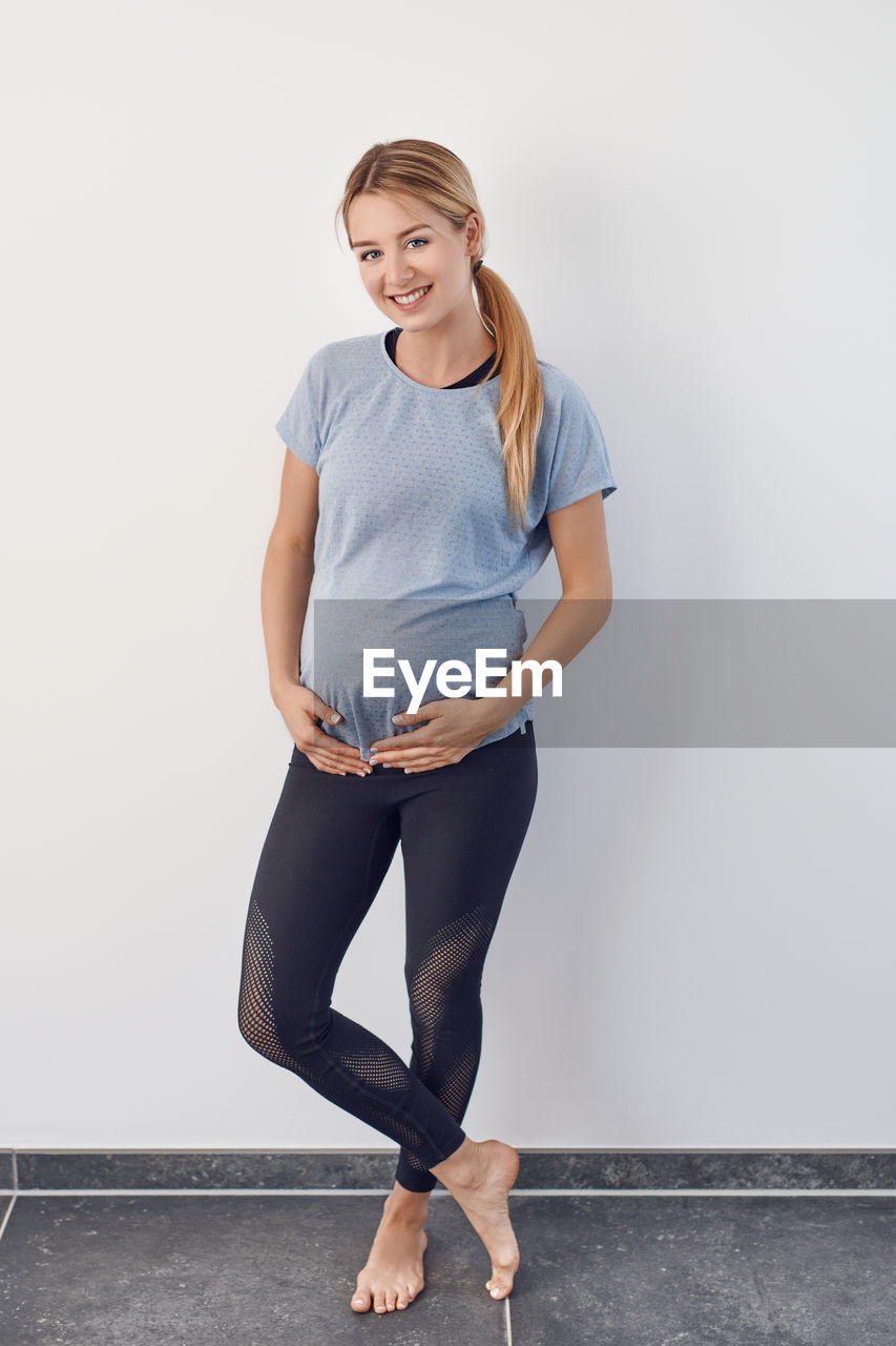 Full length portrait of smiling pregnant young woman standing against wall