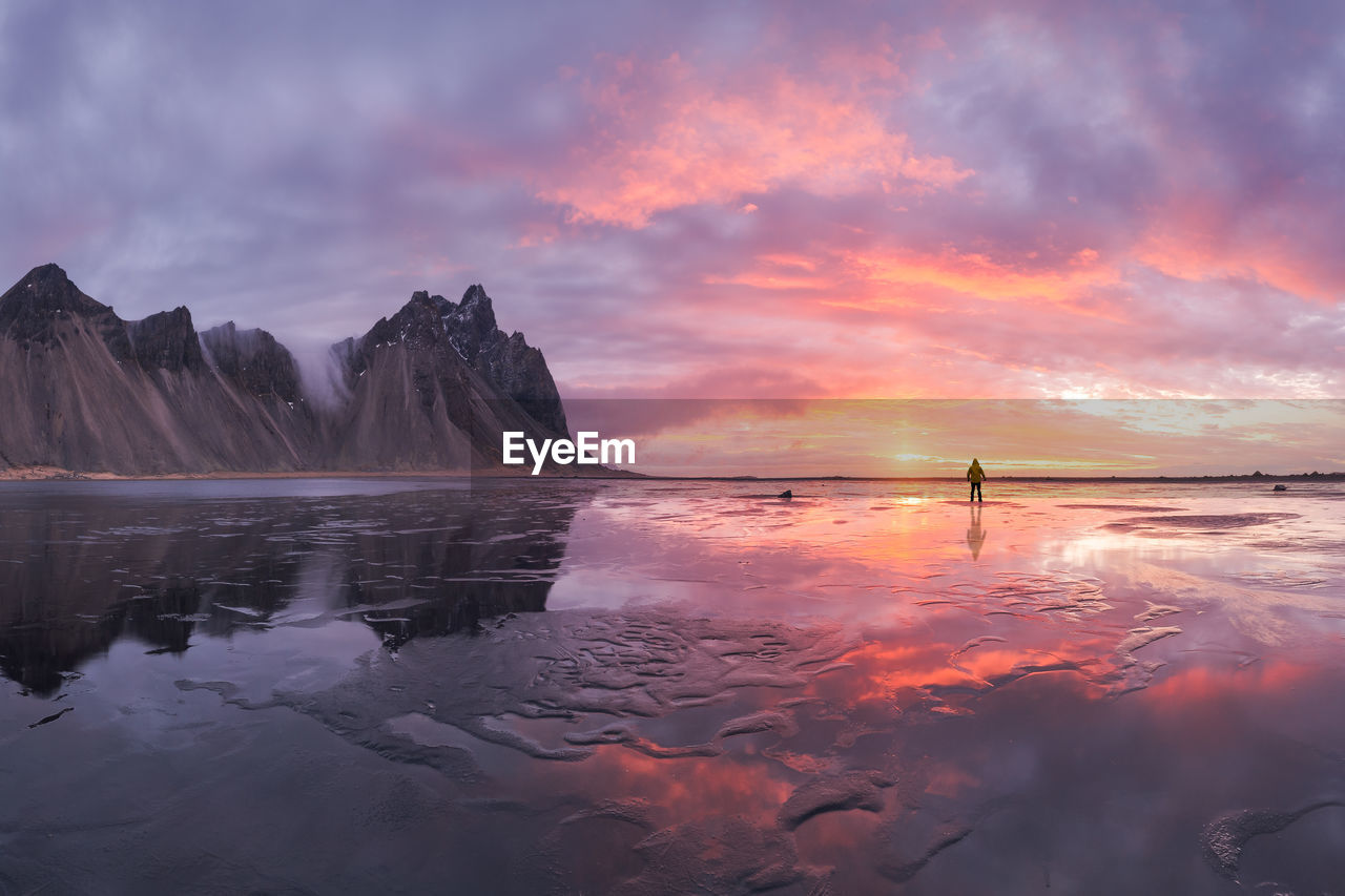 Silhouette of anonymous tourist standing on wet shore near calm sea and rough mountains during majestic colorful sunset