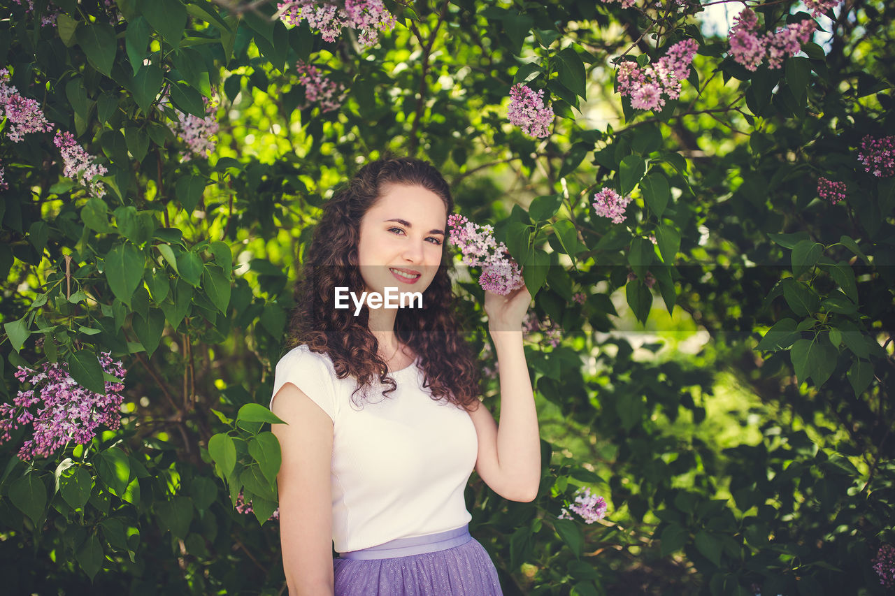 Happy curly hair young woman in a white t-shirt under the blooming lilac trees. smiling