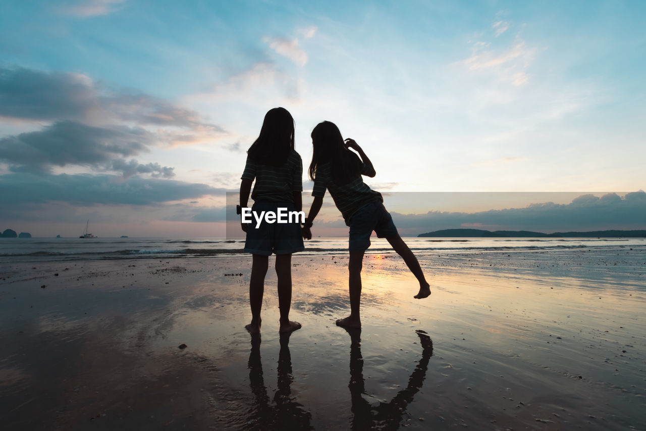 Silhouette girls holding hand standing on beach against sky